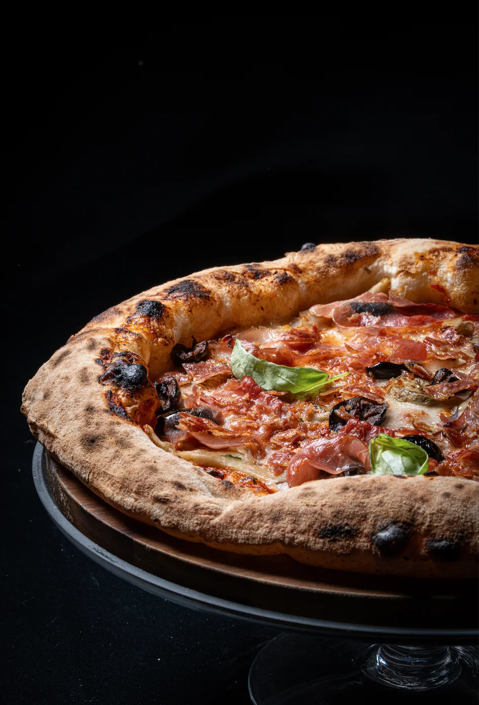 Wood-fired pizza topped with prosciutto, black olives and fresh basil on a leopard-spotted crust